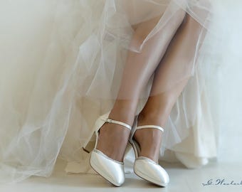 May include: White satin bridal shoes with a delicate ankle strap and a small heel. The shoes are adorned with a row of sparkling crystals.