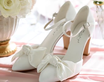 Può includere: Un paio di scarpe da sposa in raso bianco con un fiocco sulla punta. Le scarpe hanno un tacco a blocco e un cinturino alla caviglia.