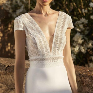 May include: A white lace wedding dress with a plunging neckline and a fitted silhouette. The dress has short sleeves and a delicate lace pattern.