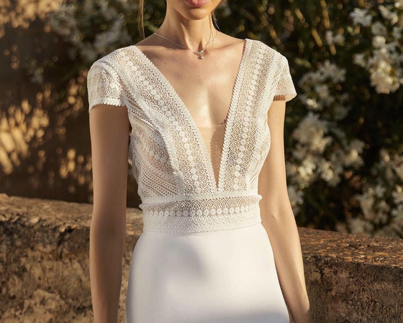 May include: A white lace wedding dress with a plunging neckline and a fitted silhouette. The dress has short sleeves and a delicate lace pattern.