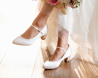 May include: A pair of white satin bridal shoes with a low heel and a strap across the foot. The shoes have a pointed toe and a simple design.