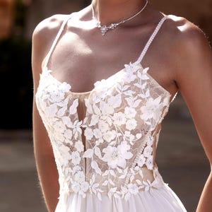 May include: A white wedding dress with a lace bodice and a flowing skirt. The bodice features delicate floral embroidery and a sheer panel in the center.