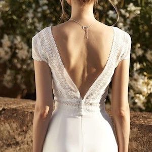 May include: A white wedding dress with a lace bodice and a deep V-back. The dress has a fitted silhouette and a flowing skirt.