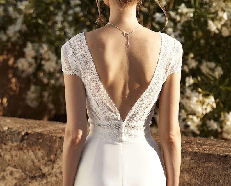 May include: A white wedding dress with a lace bodice and a deep V-back. The dress has a fitted silhouette and a flowing skirt.