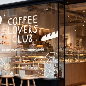 Pu&ograve; includere: Vetrina di un bar con la scritta "COFFEE LOVERS CLUB" e una selezione di pasticceria. L'orario di apertura &egrave; indicato dalle 8:00 alle 22:00. Sgabelli in legno sono posizionati davanti alla vetrina, offrendo uno sguardo all'interno.