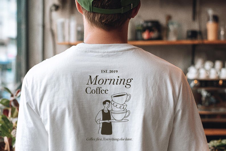 Pu&ograve; includere: T-shirt bianco con una grafica sul retro che dice "Morning Coffee" con una pila di tazze da caff&egrave; e una persona che tiene un vassoio. Il testo "Coffee first, Everything else later" &egrave; stampato anche sulla maglietta. La maglietta ha anche "EST. 2019" stampato.