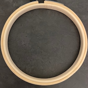 May include: A circular embroidery hoop made of light-coloured wood, featuring a metal screw and bolt for tension adjustment. The hoop is designed for holding fabric securely during embroidery or cross-stitch. The metal components are silver in colour.