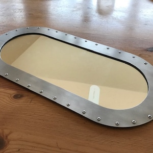 Puede incluir: Espejo de forma ovalada con un marco de metal plateado y un borde decorativo de pequeños remaches. El espejo refleja una superficie de madera marrón claro.