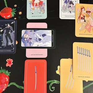 Op de afbeelding: Een verzameling tarotkaarten met kleurrijke illustraties en titels als "Queen of Wands", "Page of Wands" en "Ace of Swords". De kaarten zijn gerangschikt op een zwart oppervlak met bloemen- en fruitdecoraties.