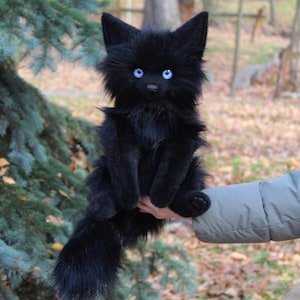 Op de afbeelding: Een zwart pluche kattenknuffel met helderblauwe ogen en een lange, pluizige staart. De kat heeft puntige oren en wordt voor een groene boom gehouden. De vacht van de kat is diepzwart.