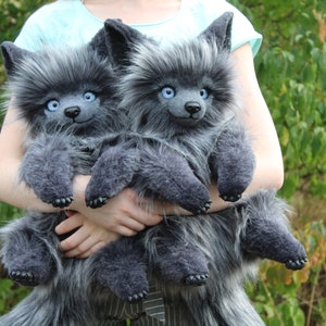 Op de afbeelding: Twee pluche wolfpoppen met grijze vacht en blauwe ogen worden voor een groene achtergrond gehouden. De poppen hebben puntige oren, kleine zwarte neuzen en donkergrijze poten. De poppen zijn zacht en pluizig.