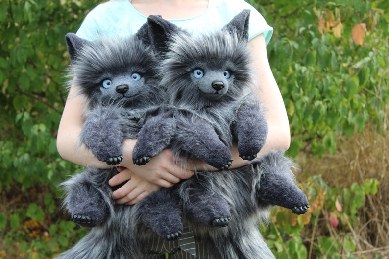 Op de afbeelding: Twee pluche wolfpoppen met grijze vacht en blauwe ogen worden voor een groene achtergrond gehouden. De poppen hebben puntige oren, kleine zwarte neuzen en donkergrijze poten. De poppen zijn zacht en pluizig.