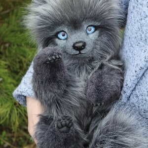 Op de afbeelding: Een zachte, grijze wolfwelp met helderblauwe ogen en een pluizige staart. Het handgemaakte knuffeldier heeft een lieve uitdrukking en wordt vastgehouden. De vacht is een mix van grijs en wit, waardoor het er realistisch uitziet.