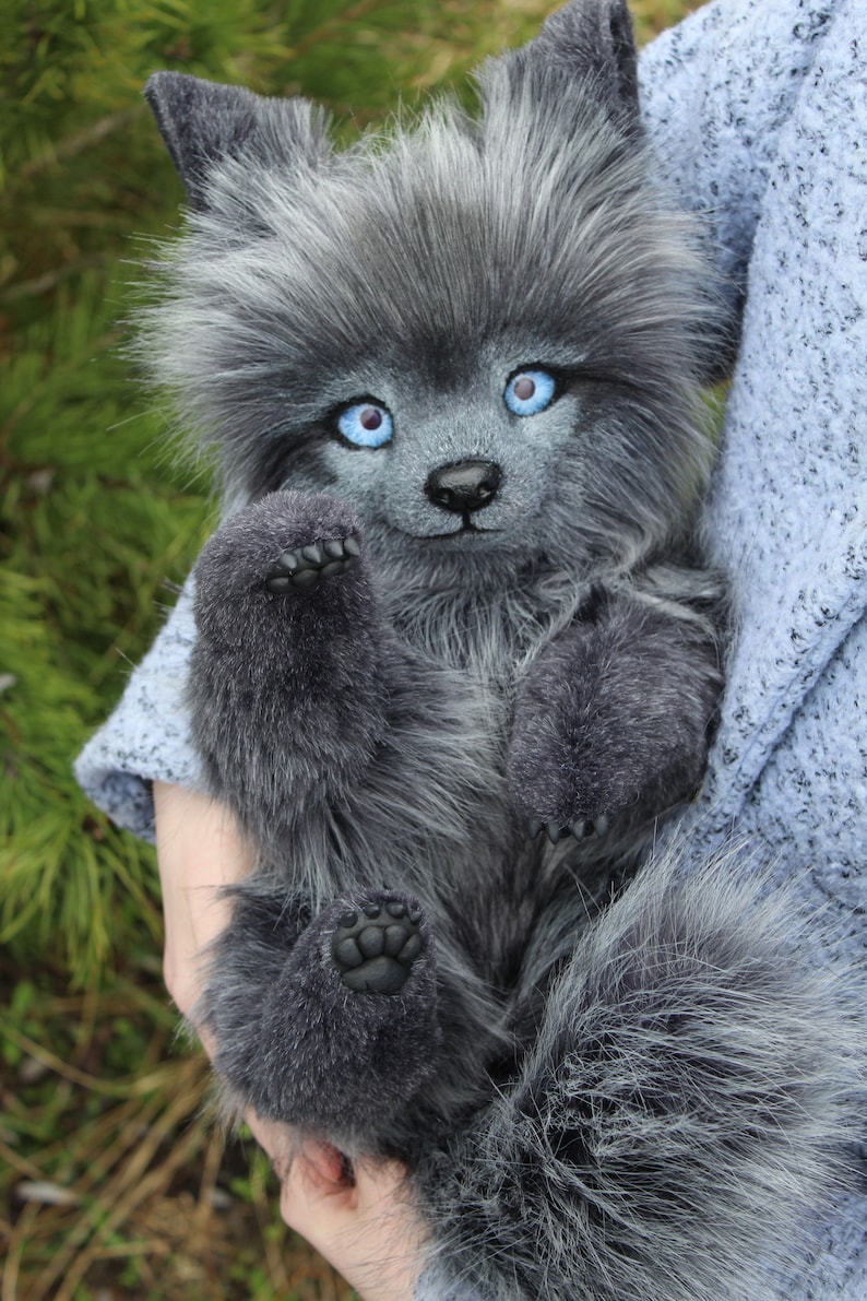 Op de afbeelding: Een zachte, grijze wolfwelp met helderblauwe ogen en een pluizige staart. Het handgemaakte knuffeldier heeft een lieve uitdrukking en wordt vastgehouden. De vacht is een mix van grijs en wit, waardoor het er realistisch uitziet.