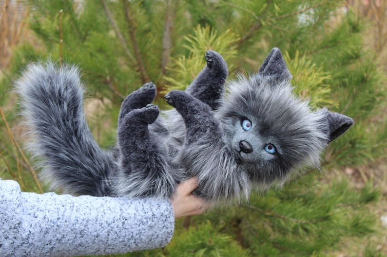 Op de afbeelding: Een handgemaakt, grijs pluche wolfwelpje met helderblauwe ogen en een pluizige staart. Het knuffeldier ligt op zijn rug, met zijn pootjes in de lucht. De achtergrond toont groene bladeren.