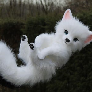 May include: A fluffy white fox plush toy with black paw pads and blue eyes. The fox has a long, bushy tail and pink inner ears. The plush is held up against a blurred green background.