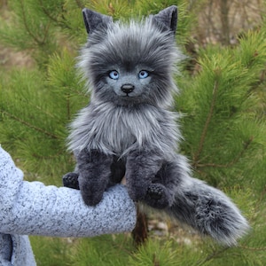 Op de afbeelding: Een grijze pluche wolf met helderblauwe ogen. Het handgemaakte dier heeft een pluizige, getextureerde vacht en zit met uitgestrekte staart. De achtergrond toont groene dennennaalden.