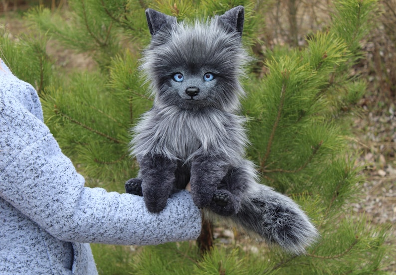 Op de afbeelding: Een grijze pluche wolf met helderblauwe ogen. Het handgemaakte dier heeft een pluizige, getextureerde vacht en zit met uitgestrekte staart. De achtergrond toont groene dennennaalden.