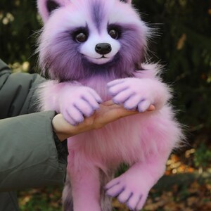 Puede incluir: Un mu&ntilde;eco de mapache de peluche rosa y morado con una textura de pelaje esponjoso. El mapache tiene una cara blanca, una nariz negra y ojos oscuros. La mu&ntilde;eca est&aacute; siendo sostenida por una persona.
