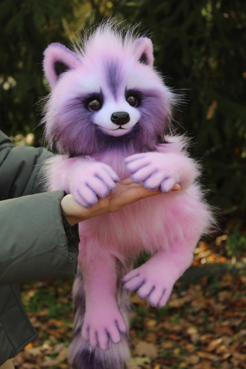 Puede incluir: Un mu&ntilde;eco de mapache de peluche rosa y morado con una textura de pelaje esponjoso. El mapache tiene una cara blanca, una nariz negra y ojos oscuros. La mu&ntilde;eca est&aacute; siendo sostenida por una persona.