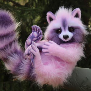 Puede incluir: Un peluche de mapache en tonos rosas y morados. El mapache tiene una cola rayada morada, un cuerpo rosa y una cara blanca y morada. Est&aacute; levantando sus patas, con una nariz negra y ojos grandes y oscuros.