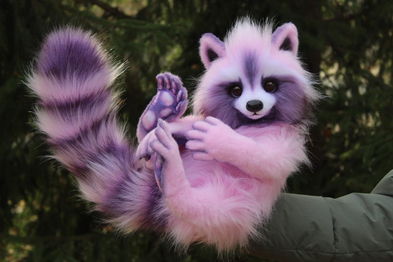 Puede incluir: Un peluche de mapache en tonos rosas y morados. El mapache tiene una cola rayada morada, un cuerpo rosa y una cara blanca y morada. Est&aacute; levantando sus patas, con una nariz negra y ojos grandes y oscuros.