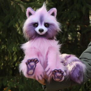 Puede incluir: Un peluche de mapache sentado en tonos rosas y morados. El juguete tiene un aspecto esponjoso y texturizado, con una cara blanca, ojos oscuros y una nariz negra. Las patas y la cola del mapache son de un morado m&aacute;s oscuro, sobre un fondo verde borroso.