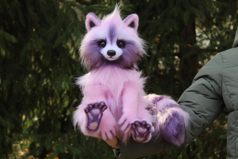 Puede incluir: Un peluche de mapache sentado en tonos rosas y morados. El juguete tiene un aspecto esponjoso y texturizado, con una cara blanca, ojos oscuros y una nariz negra. Las patas y la cola del mapache son de un morado m&aacute;s oscuro, sobre un fondo verde borroso.