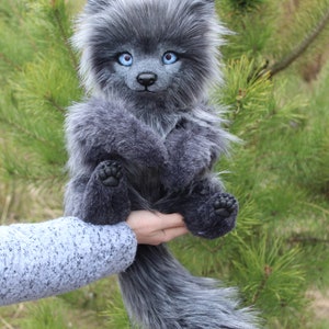 Op de afbeelding: Een grijze wolf knuffel met helderblauwe ogen en een lange, pluizige staart. Het handgemaakte knuffeldier heeft gedetailleerde kenmerken en wordt tegen een achtergrond van groen gebladerte gehouden.