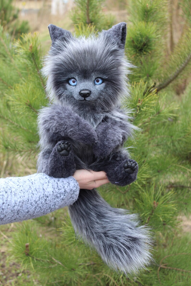 Op de afbeelding: Een grijze wolf knuffel met helderblauwe ogen en een lange, pluizige staart. Het handgemaakte knuffeldier heeft gedetailleerde kenmerken en wordt tegen een achtergrond van groen gebladerte gehouden.