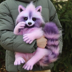Puede incluir: Un peluche de mapache lavanda y morado con una cola y patas esponjosas. El mapache tiene una cara blanca y una nariz negra. El peluche est&aacute; siendo sostenido por una persona que lleva una chaqueta verde.