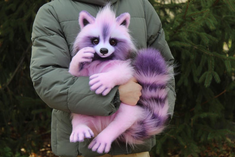 Puede incluir: Un peluche de mapache lavanda y morado con una cola y patas esponjosas. El mapache tiene una cara blanca y una nariz negra. El peluche est&aacute; siendo sostenido por una persona que lleva una chaqueta verde.