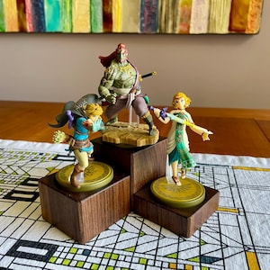 Op de afbeelding: Drie karakterbeeldjes uit de Legend of Zelda-franchise worden tentoongesteld op een houten trapstandaard. De figuren zijn onder meer Link, Ganon en Zelda, elk met gedetailleerde kostuums en accessoires. De standaard staat op een tafelkleed met patroon.