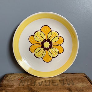 以下が含まれることがあります： 黄色の縁取りと中央に茶色と黄色の花柄が描かれた白いセラミック製のプレートです。