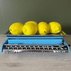 May include: A vintage blue and silver kitchen scale with a tray holding five yellow lemons. The scale has a black dial with white markings and the text "Marianne". The scale measures up to 12 kg.