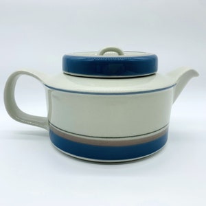 Op de afbeelding: Een witte theepot van keramiek met een blauw en bruin gestreept ontwerp. De theepot heeft een deksel met een knop aan de bovenkant.