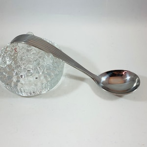 Könnte beinhalten: Ein Edelstahl-Löffel mit geriffeltem Griff und abgerundeter Schale. Der Löffel liegt auf einem transparenten, strukturierten Glasobjekt. Der Löffel hat eine glänzende silberne Farbe.