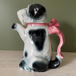 Könnte beinhalten: Eine keramische Teekanne in Hundeform mit einem schwarz-weiß gefleckten Körper, einer rosa Schleife und einem rosa Griff. Die Teekanne hat einen rosa Kragen und einen Deckel, der den Kopf des Hundes bildet. Die Teekanne steht auf einer hellen Oberfläche.