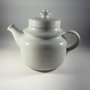 Op de afbeelding: Witte theepot van keramiek met een deksel en een handvat. De theepot heeft een glad, rond lichaam en een licht uitlopende tuit.