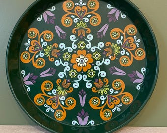 May include: A round, dark green metal tray with a vibrant floral design. The pattern features orange, yellow, purple, and white stylized flowers and leaves. The tray's rim is a solid dark green.