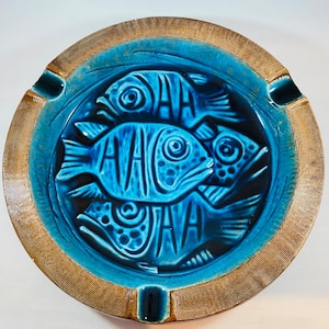 May include: A blue ceramic ashtray with a brown rim. The ashtray features a design of three stylized fish in a blue and white pattern.