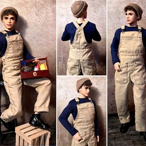 May include: A doll wearing a brown beanie and a beige overall with a blue long-sleeved shirt underneath. The doll is holding a toolbox with tools inside.