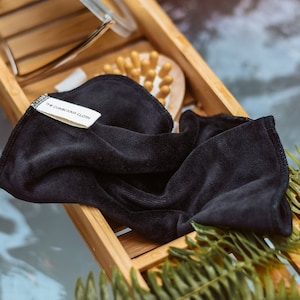 Peut inclure: Un gant de toilette noir avec le texte "The Conscious Co." sur un plateau en bois avec une foug&egrave;re verte. Le plateau est dans une baignoire avec de l'eau.