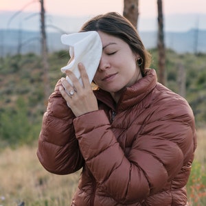Könnte beinhalten: Eine Frau in einer braunen Daunenjacke und schwarzen Hosen sitzt auf einem Baumstamm und wischt sich mit einem weißen Tuch die Stirn. Sie ist im Freien in einem grasbewachsenen Gebiet mit Bäumen im Hintergrund.