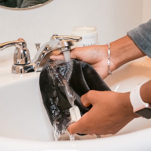 Peut inclure: Une personne rince un masque de sommeil en velours noir sous l'eau courante dans un &eacute;vier en porcelaine blanche. Le masque de sommeil a une &eacute;tiquette blanche avec le mot "SLEEP" imprim&eacute; dessus.