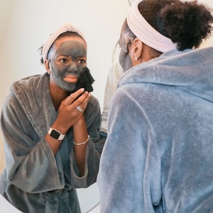 Peut inclure: Une personne portant une robe grise et un masque facial noir applique le masque sur son visage tout en regardant dans un miroir.