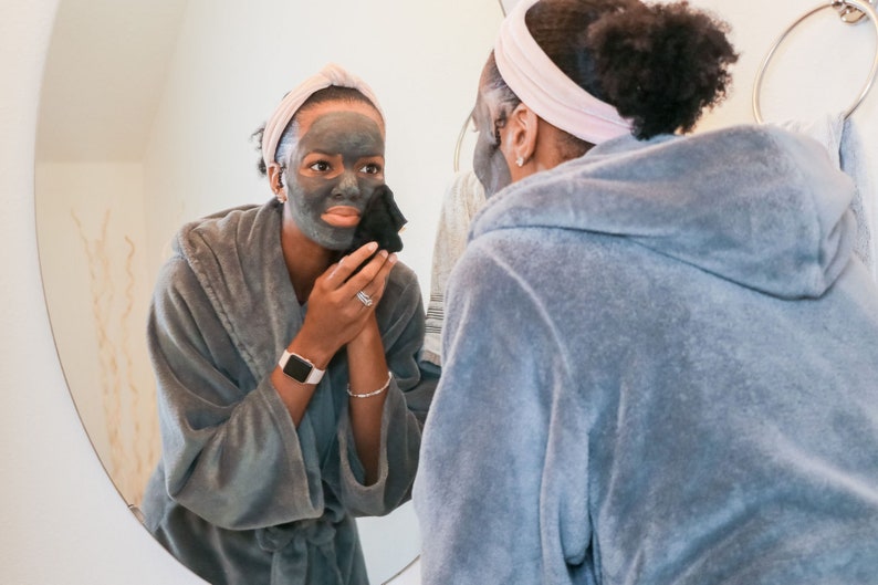 Peut inclure: Une personne portant une robe grise et un masque facial noir applique le masque sur son visage tout en regardant dans un miroir.