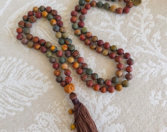 May include: A long beaded necklace featuring matte, multi-colored stone beads in shades of red, green, and yellow. The necklace includes several textured, carved wooden accent beads, a dark brown fabric tassel, and a small circular metal charm.