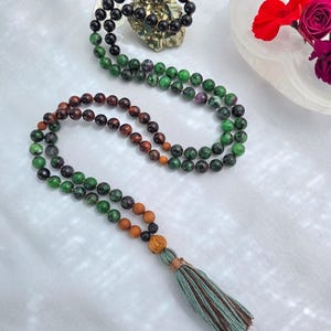 Könnte beinhalten: Zwei Perlenketten mit Quasten. Eine Halskette hat grüne und braune Perlen, während die andere schwarze und grüne Perlen hat. Die Halsketten werden auf einer weißen Oberfläche präsentiert, mit einer kleinen Schale mit Blumen und einem Kristall im Hintergrund.