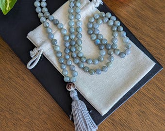 May include: A grey gemstone mala necklace with a tassel, resting on a white cloth pouch and a black surface. The necklace is made of round beads and has a brown wooden bead at the end.